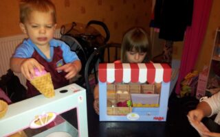 A young boy holds a toy ice cream cone beside a toy ice cream set, while a young girl plays with a pretend fruit stall. Another person’s hand appears on the right, possibly handling toy money. The setting appears to be a playroom or dining area.