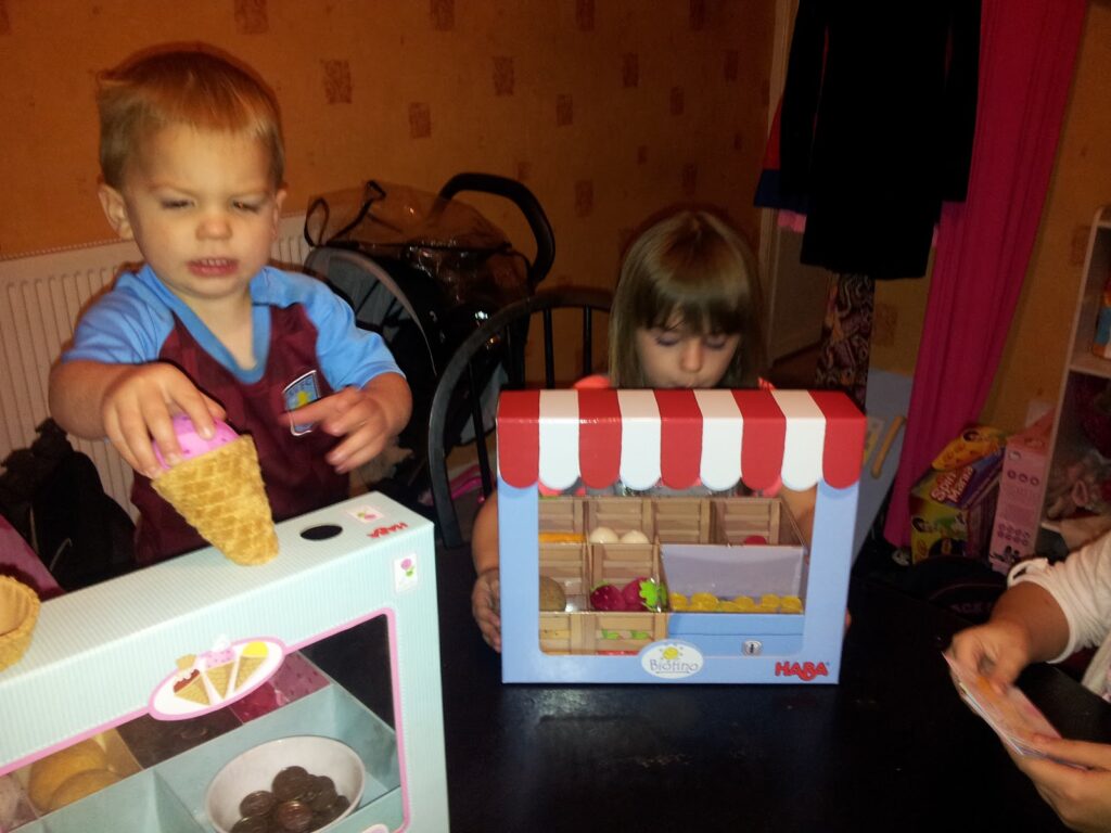 Two young children play at a table with toy food sets. The boy, wearing a football shirt, places a scoop on a toy ice cream cone, while the girl sits beside a toy market stall with play fruit. An adult’s hand with cards is visible at the right edge.