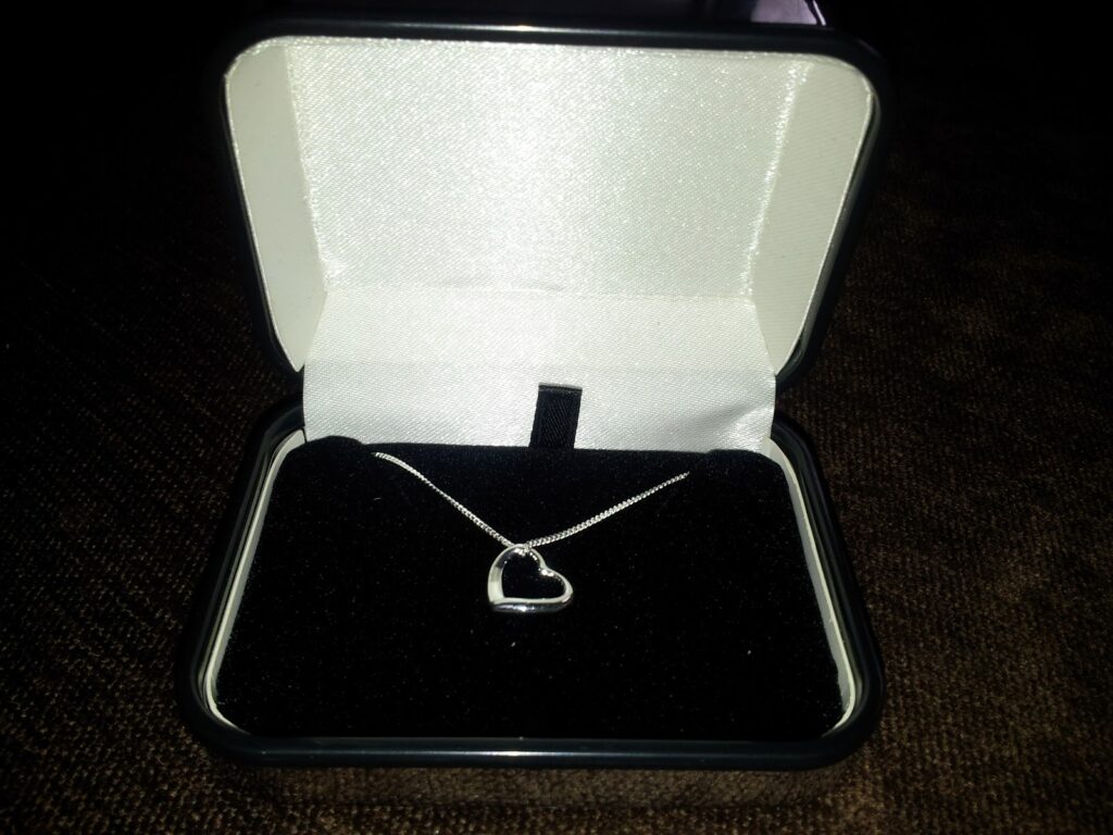 A silver heart-shaped pendant on a fine chain is displayed in an open black and white jewellery box with a soft black velvet interior, sitting on a dark textured surface. The box lid is lined with white satin.