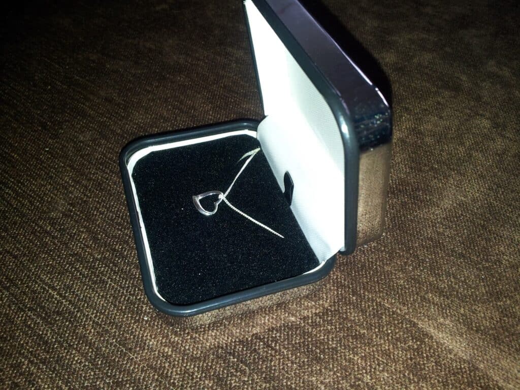 A silver heart-shaped pendant necklace sits in a black velvet jewellery box with a white interior. The box is open and placed on a brown textured surface. The pendant rests on the black lining, and the chain is slightly coiled beside it.