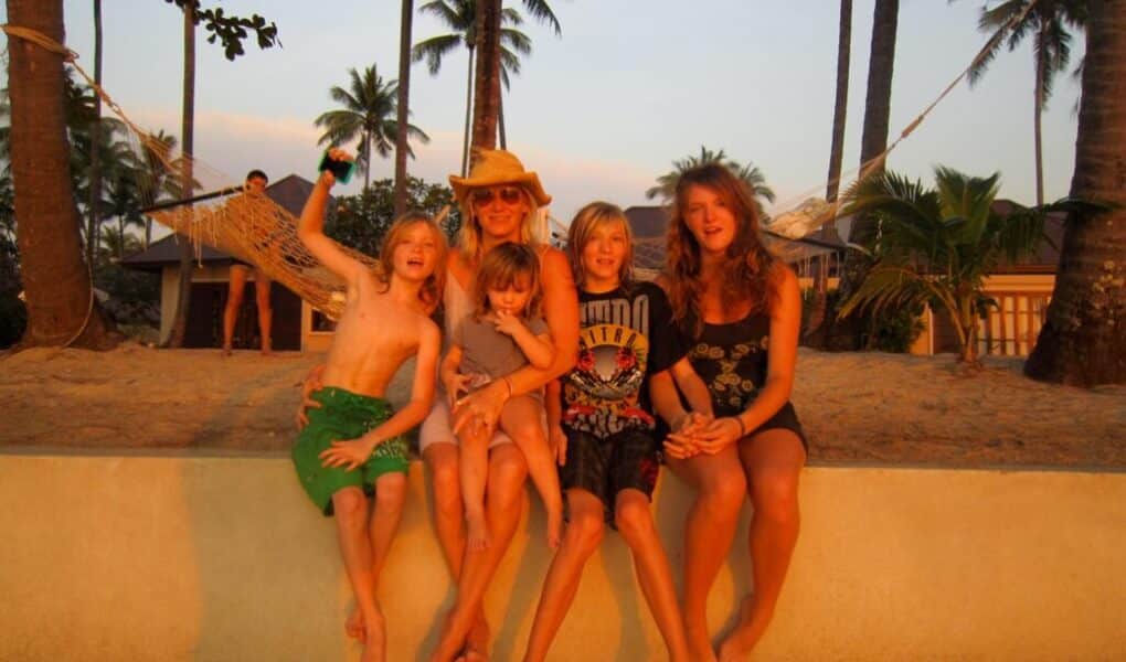 A woman in a sunhat sits on a low wall at the beach with four children, all smiling and barefoot. Palm trees and a hammock are in the background, and the group is bathed in warm sunset light. The mood is joyful and relaxed.