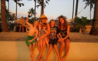 A woman in a sunhat sits on a low wall at the beach with four children, all smiling and barefoot. Palm trees and a hammock are in the background, and the group is bathed in warm sunset light. The mood is joyful and relaxed.