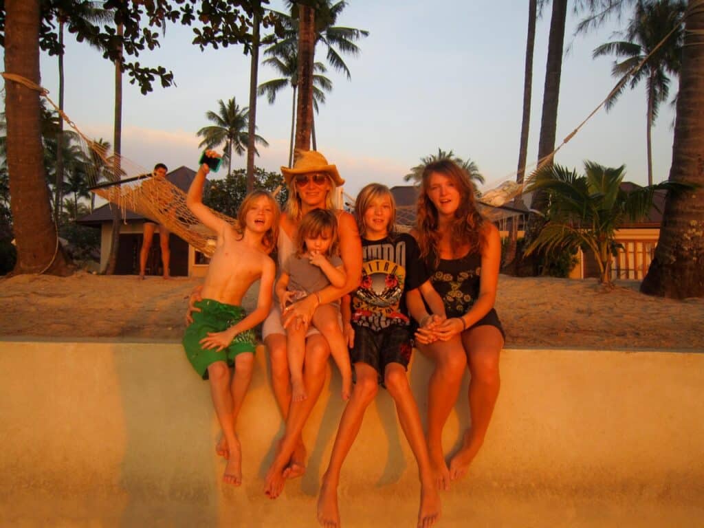 A woman in a sunhat sits on a ledge with four smiling children in swimsuits, all bathed in warm sunlight. Palm trees, a hammock, and a person standing in the background suggest a tropical beach setting at sunset.