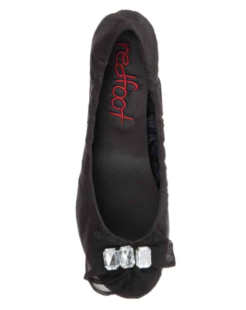 A black ballet flat shoe viewed from above, featuring a decorative bow with three large clear gemstones on the toe. The insole is black with "redfoot" written in red text near the heel. The shoe appears soft and has a velvet-like texture.