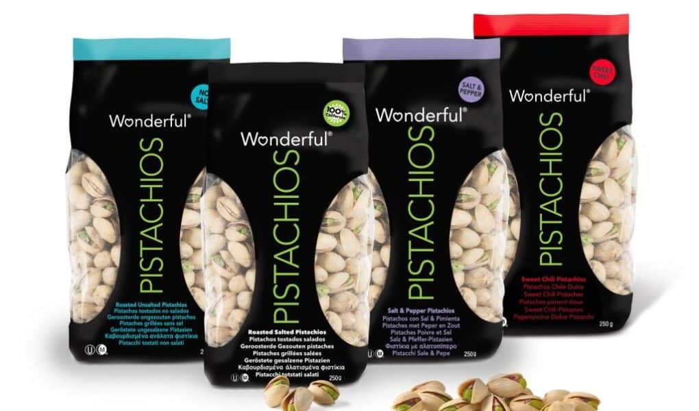 Four colourful bags of Wonderful Pistachios—Roasted Unsalted, Roasted Salted, Salt & Pepper, and Sweet Chilli—stand upright. Each bag has a black centre with clear side panels showing pistachios inside. A few shelled pistachios lie in front.