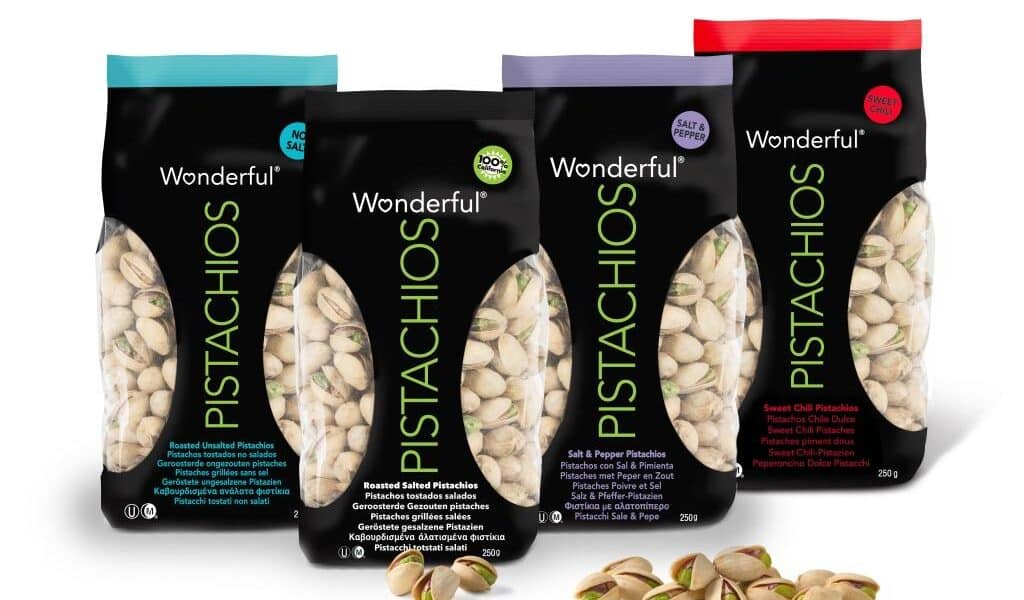 Four colourful bags of Wonderful Pistachios—Roasted Unsalted, Roasted Salted, Salt & Pepper, and Sweet Chilli—stand upright. Each bag has a black centre with clear side panels showing pistachios inside. A few shelled pistachios lie in front.