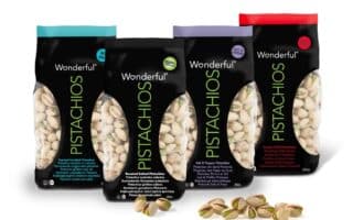 Four colourful bags of Wonderful Pistachios—Roasted Unsalted, Roasted Salted, Salt & Pepper, and Sweet Chilli—stand upright. Each bag has a black centre with clear side panels showing pistachios inside. A few shelled pistachios lie in front.