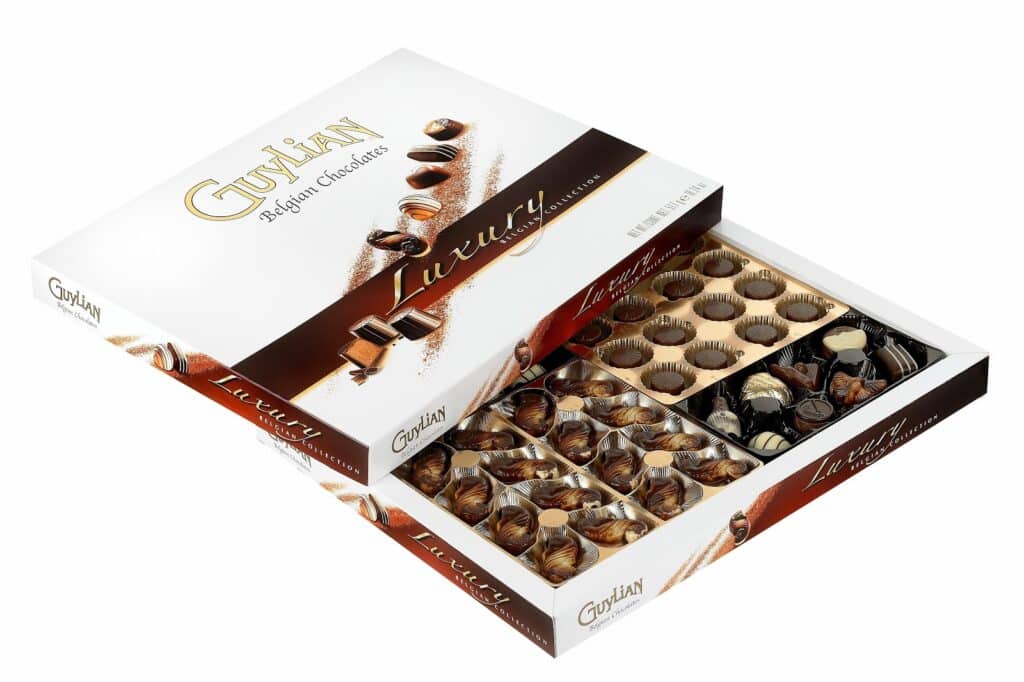 An open box of Guylian Belgian Chocolates shows assorted seashell-shaped chocolates arranged neatly in plastic trays. The box lid leans against the back, revealing "Guylian Luxury" branding with images of chocolates on a white and brown background.