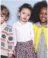 Three young children stand side by side. The child on the left wears a patterned jumper, the child in the middle wears a white shirt with a floral skirt, and the child on the right smiles in a grey dress with a yellow cardigan. They pose against a light blue background.