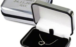 A silver jewellery box with a white interior holds a delicate heart-shaped necklace. The lid, propped open, reveals an engraved message: "With my love... today, tomorrow and always! 24.11.2011 XXX." Another closed box shows the engraving clearly.