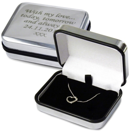 A silver jewellery box with a white interior holds a delicate heart-shaped necklace. The lid, propped open, reveals an engraved message: "With my love... today, tomorrow and always! 24.11.2011 XXX." Another closed box shows the engraving clearly.