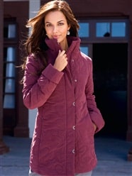 A woman with long brown hair stands outside in front of a building, wearing a mauve quilted coat with a high collar. She is holding the collar with one hand and smiling softly, looking confidently at the camera. The background shows large windows and a doorway.