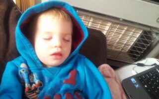 A young child with blond hair is sleeping whilst sitting up, wearing a blue hoodie with Thomas the Tank Engine on it. The child’s lips are slightly parted. Nearby is a black laptop and a heater in the background. The scene looks cosy and peaceful.