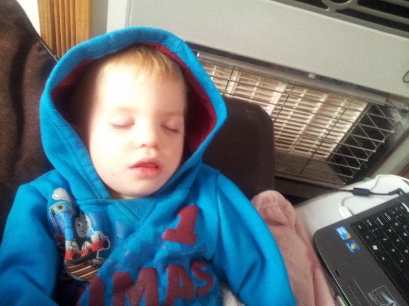 A young child with blond hair is sleeping whilst sitting up, wearing a blue hoodie with Thomas the Tank Engine on it. The child’s lips are slightly parted. Nearby is a black laptop and a heater in the background. The scene looks cosy and peaceful.