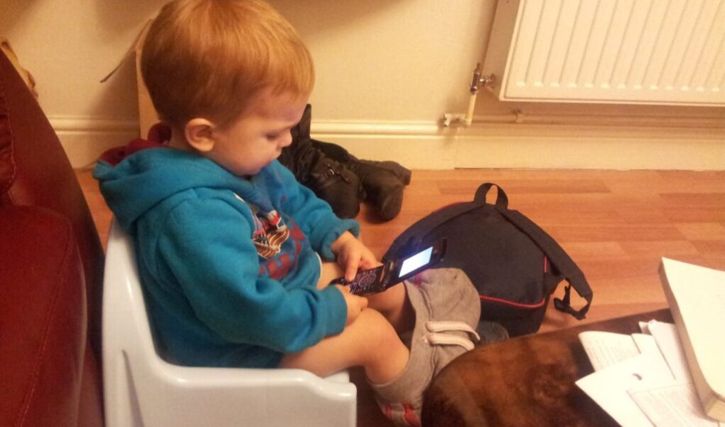A young child with short red hair sits on a light blue plastic chair indoors, wearing a blue hoodie and grey trousers. The child is focused on an open flip phone, with a dark rucksack, shoes, and scattered papers nearby on the floor and table.