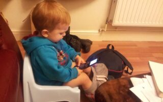 A young child with short red hair sits on a light blue plastic chair indoors, wearing a blue hoodie and grey trousers. The child is focused on an open flip phone, with a dark rucksack, shoes, and scattered papers nearby on the floor and table.
