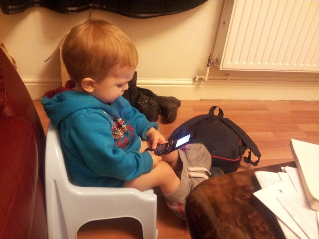 A young child with light brown hair, wearing a blue hoodie and grey shorts, sits on a plastic chair. The child is focused on holding and pressing buttons on a flip phone. There are papers, a bag, and shoes around on the wooden floor.