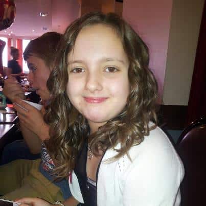 A young girl with long, curly brown hair and a white cardigan smiles at the camera in a restaurant. In the background, a boy in a grey shirt is eating, holding a spoon and looking to the side. The setting is warmly lit with red and white decor.