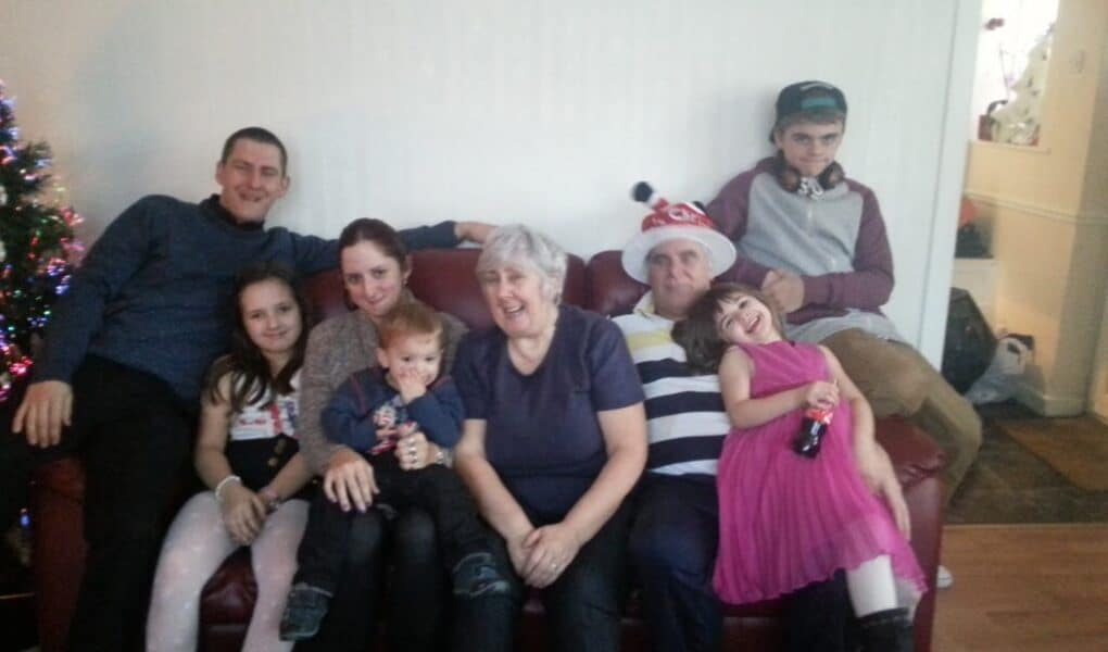A family of seven sits closely on a red sofa. An older woman and man sit in the centre, surrounded by four children and two adults. The man wears a striped shirt and festive hat. Everyone is smiling, and a Christmas tree is partially visible on the left.