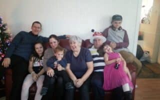 A family of seven sits closely on a red sofa. An older woman and man sit in the centre, surrounded by four children and two adults. The man wears a striped shirt and festive hat. Everyone is smiling, and a Christmas tree is partially visible on the left.