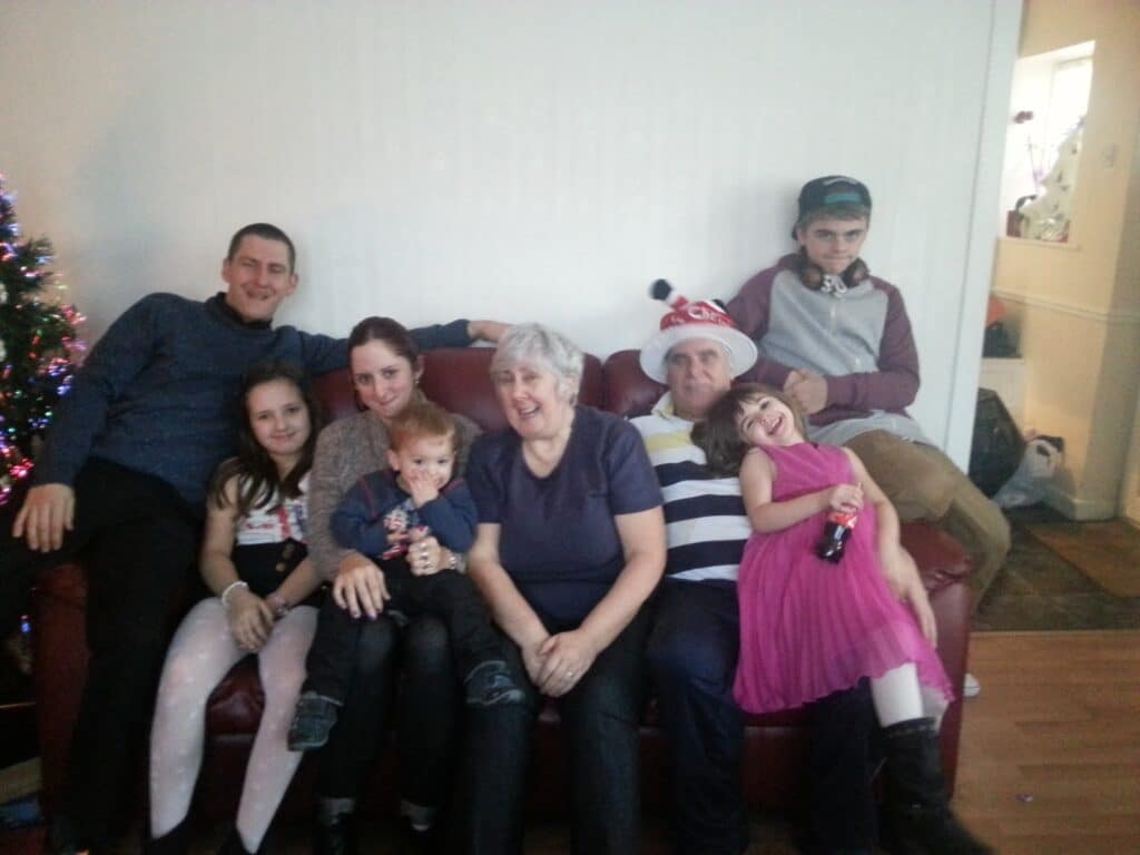 Seven people sit and pose on a red sofa in a living room. Three adults sit in the centre, surrounded by four children of various ages. A Christmas tree is partially visible on the left, and one person wears a festive Father Christmas hat. Everyone appears happy and relaxed.