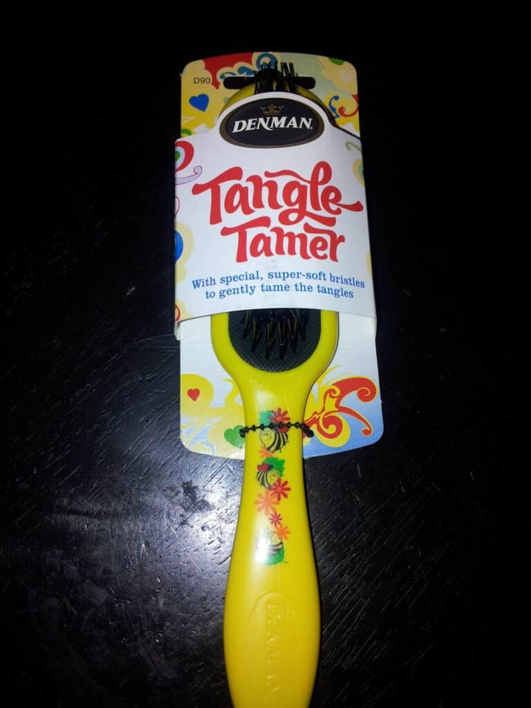 A Denman Tangle Tamer hairbrush in yellow packaging with decorative hearts and flowers. The label reads, "Tangle Tamer: With special, super-soft bristles to gently tame the tangles." The brush handle has a floral pattern.