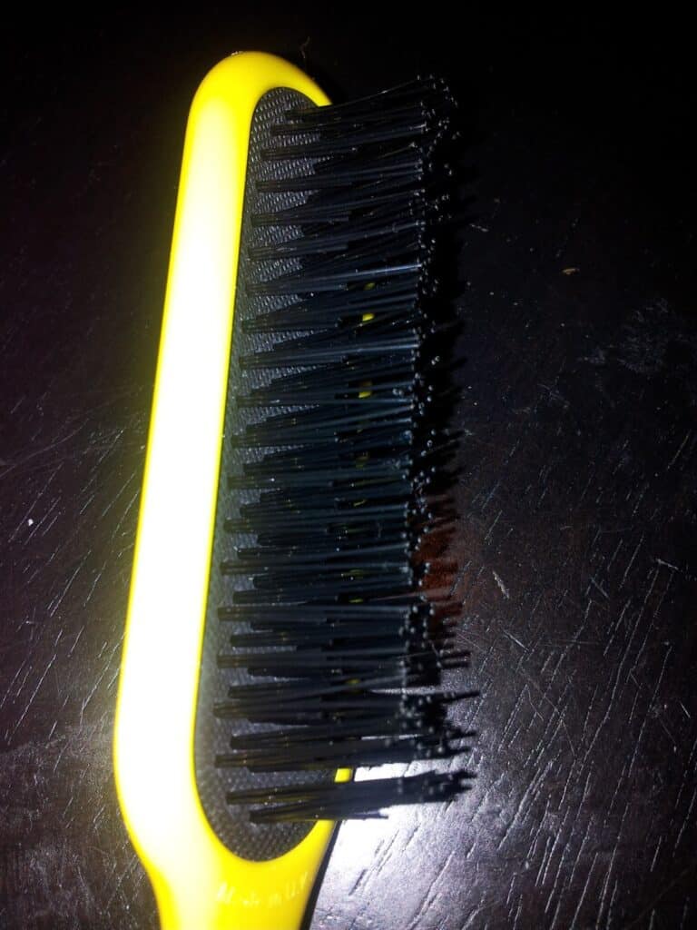 A close-up photo of a yellow hairbrush with black plastic bristles, shown against a dark, scratched surface. The brush is slightly angled, and the bristles appear clean and shiny.
