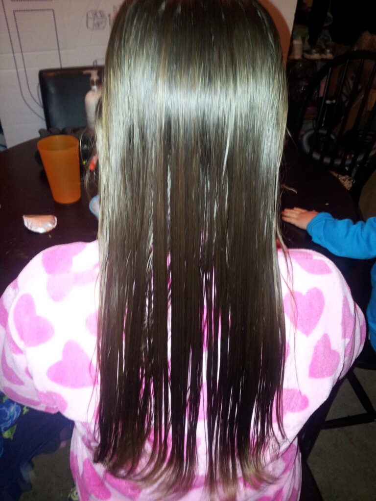 A child with long, straight, wet brown hair is shown from behind. The child is wearing a white dressing gown with large pink hearts. The background includes a dark table with an orange cup, a white wall, and partially visible people.