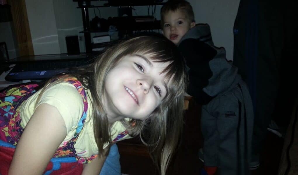 A young girl with long brown hair, wearing a colourful dress over a yellow shirt, smiles and leans forward towards the camera. Behind her, a small boy in grey trousers and a black hoodie looks on. The scene appears to be indoors, near a TV stand.