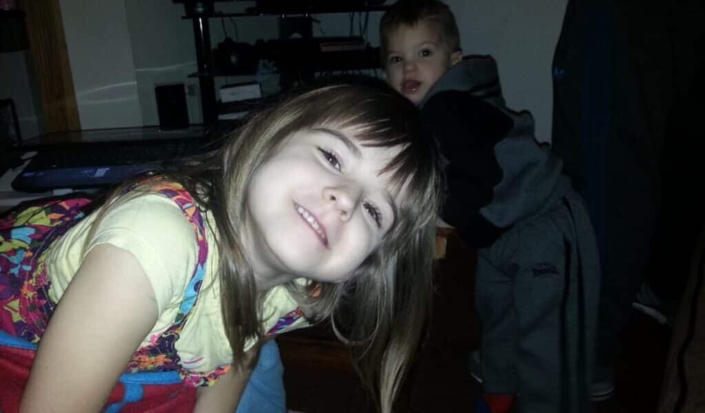 A young girl with long brown hair, wearing a colourful dress over a yellow shirt, smiles and leans forward towards the camera. Behind her, a small boy in grey trousers and a black hoodie looks on. The scene appears to be indoors, near a TV stand.
