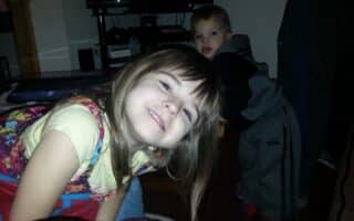 A young girl with long brown hair, wearing a colourful dress over a yellow shirt, smiles and leans forward towards the camera. Behind her, a small boy in grey trousers and a black hoodie looks on. The scene appears to be indoors, near a TV stand.