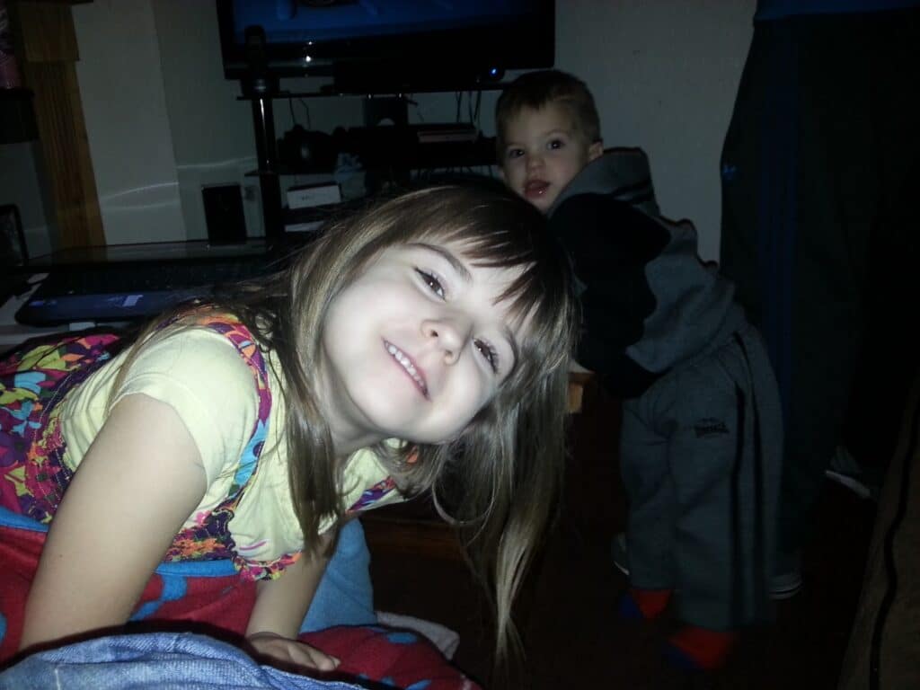 A young girl with brown hair and a floral dress smiles at the camera, leaning forward. Behind her, a small boy in a grey hoodie and tracksuit bottoms looks towards the camera. An adult, mostly out of shot, stands nearby. The room is dimly lit, with a telly in the background.