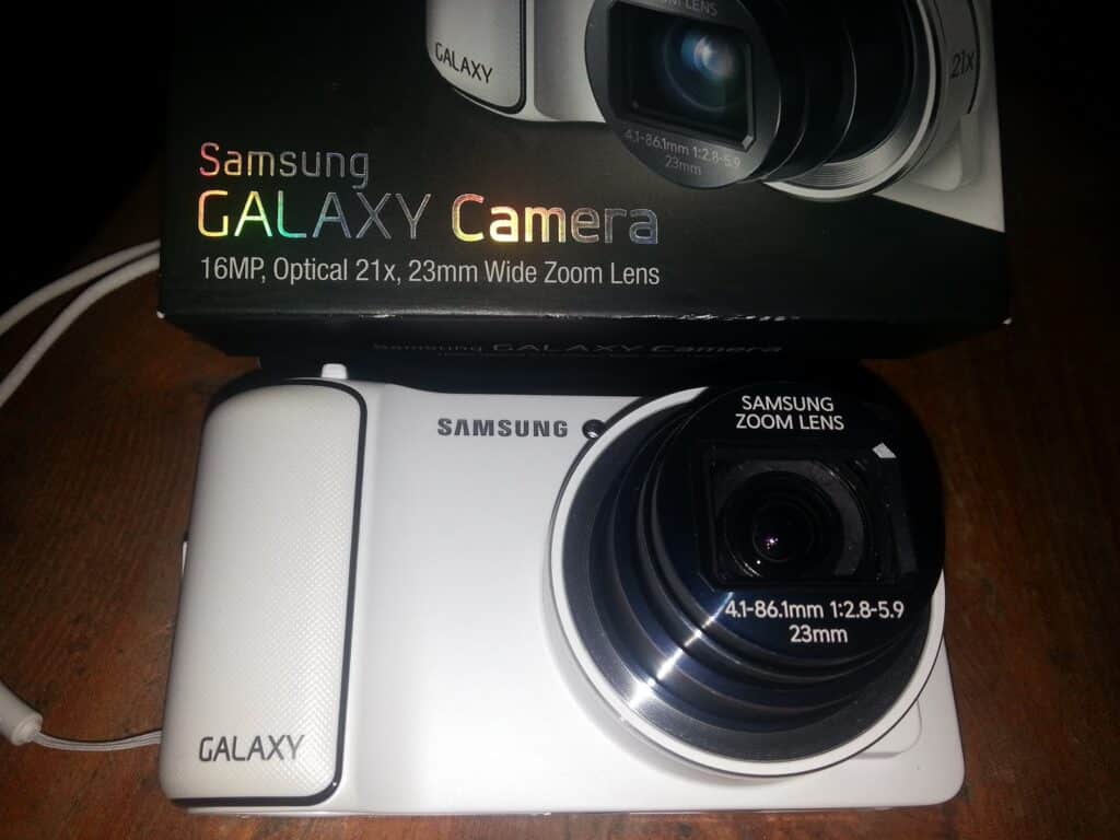 A white Samsung Galaxy Camera with a large lens is shown on a wooden surface. The lens displays "SAMSUNG ZOOM LENS 4.1-86.1mm 1:2.8-5.9 23mm." Behind it is the product box with the text "Samsung GALAXY Camera 16MP, Optical 21x, 23mm Wide Zoom Lens.