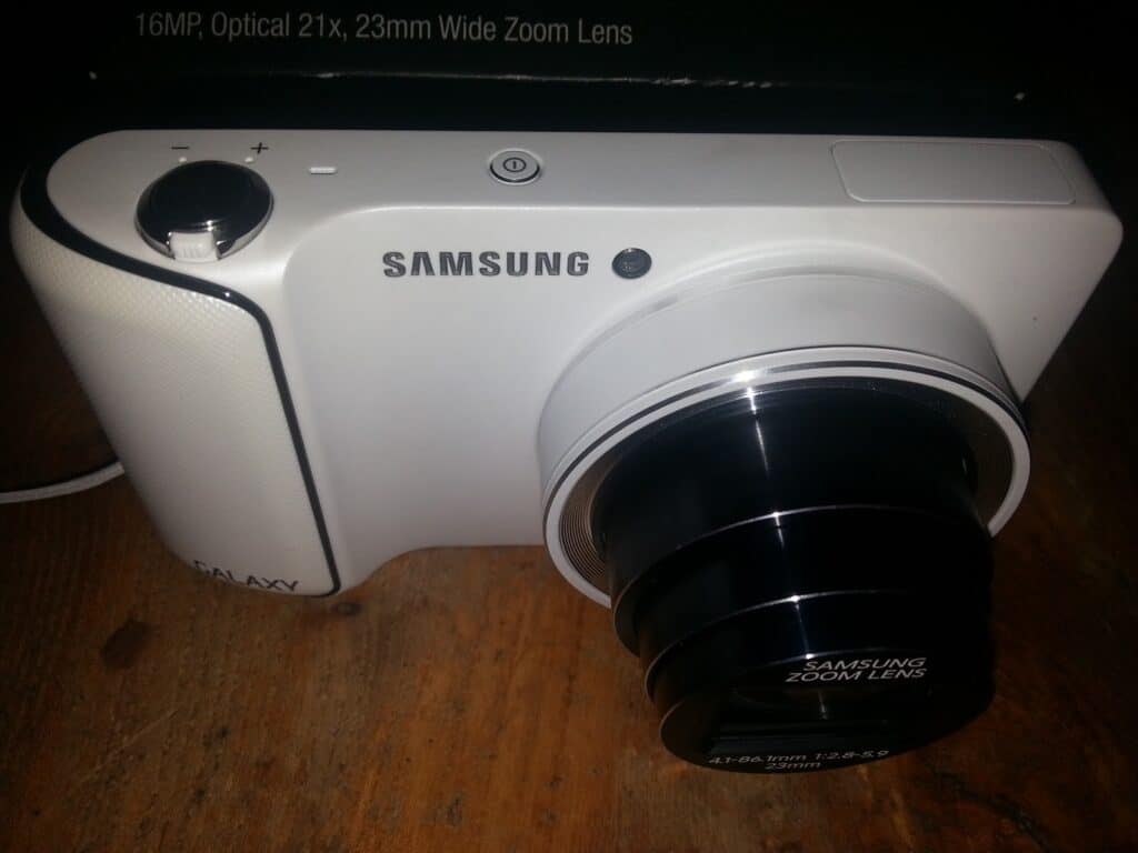 A white Samsung digital camera with a black Samsung Zoom Lens is placed on a wooden surface. “16MP, Optical 21x, 23mm Wide Zoom Lens” is written at the top of the image. The camera is angled to show its lens and top controls.