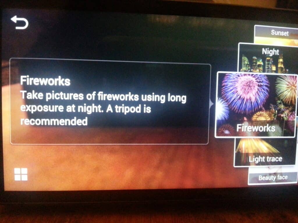 A camera screen displays shooting modes. The "Fireworks" mode is selected, showing an image of colourful fireworks. The instructions read: "Take pictures of fireworks using long exposure at night. A tripod is recommended." Other options include Night and Light trace.
