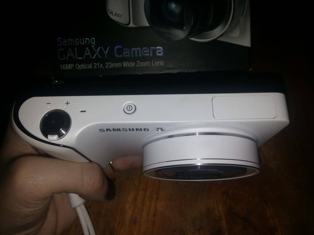 A hand holds a white Samsung Galaxy Camera, showing the top view with a control dial, power button, and lens. The camera box is in the background, displaying the model name and lens specifications: 16MP, Optical 21x, 23mm Wide Zoom Lens, on a wooden surface.