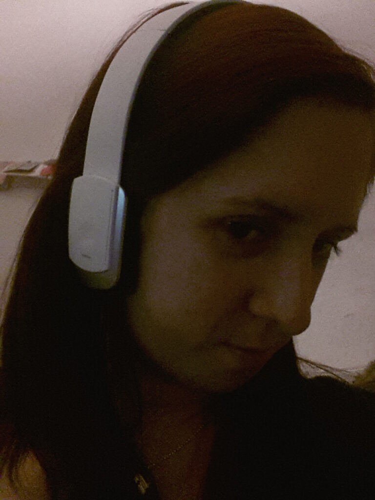 A person with long dark hair is wearing white over-ear headphones and a dark top, looking slightly downward and towards the camera. The lighting is dim and the background is blurry, with indistinct objects visible on a shelf.
