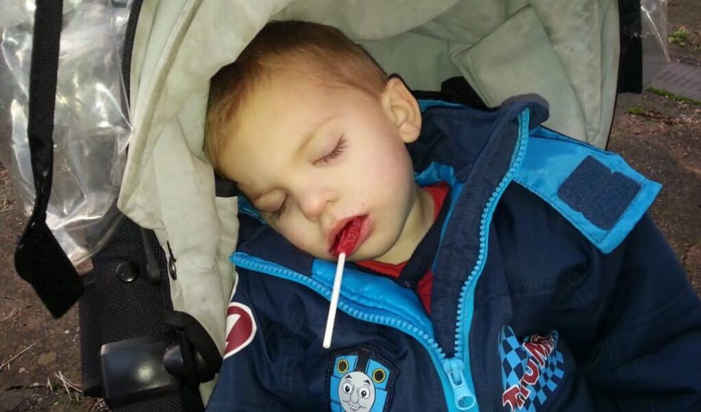 A young child in a blue jacket with a Thomas the Tank Engine patch sleeps in a pushchair, with eyes closed and a lolly in their mouth. The pushchair has a light-coloured hood, and the child looks peaceful and comfortable.