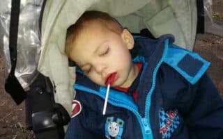 A young child in a blue jacket with a Thomas the Tank Engine patch sleeps in a pushchair, with eyes closed and a lolly in their mouth. The pushchair has a light-coloured hood, and the child looks peaceful and comfortable.