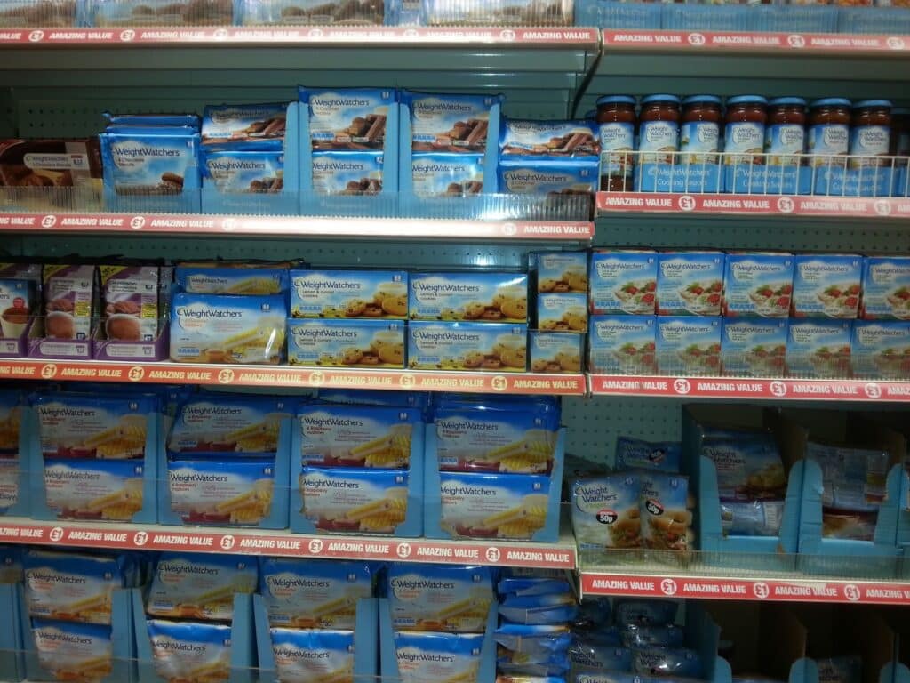 Shelves in a supermarket are stocked with Weight Watchers branded frozen meals and desserts in blue packaging, including ready meals, snack bars, and desserts. A row of blue jars is visible on the top right shelf. Red shelf labels read "AMAZING VALUE.