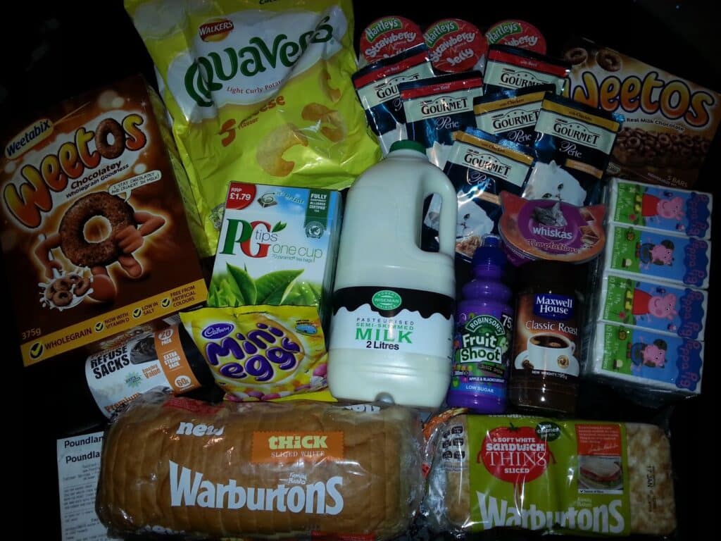 A variety of groceries on a black background, including milk, bread, Weetabix Oateos cereal, Quavers, Fridge Raiders, Mini Eggs, Fruit Shoot drink, Whiskas cat food, Maxwell House coffee, Petits Filous yoghurts, and Heinz sandwich spread.