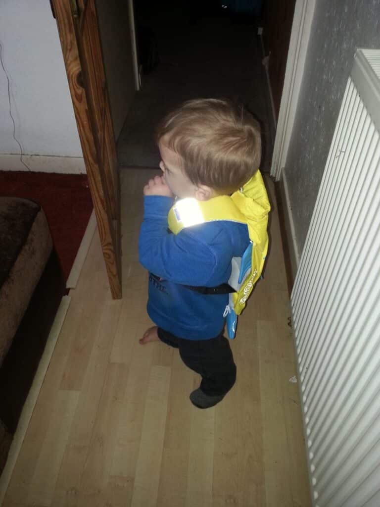 A young child wearing a blue shirt, dark trousers, and a yellow flotation device stands indoors on a light wooden floor beside a white radiator, facing a partially open wooden door. The child is sucking their thumb and looking away from the camera.