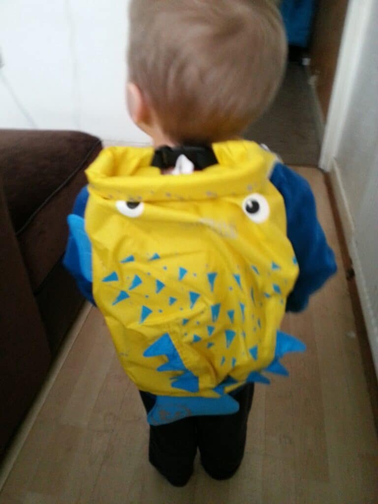 A small child with light brown hair, seen from behind, wears a bright yellow rucksack decorated with blue triangles and cartoonish eyes, resembling a playful fish or monster. The child stands on a wooden floor in a corridor next to a brown sofa.