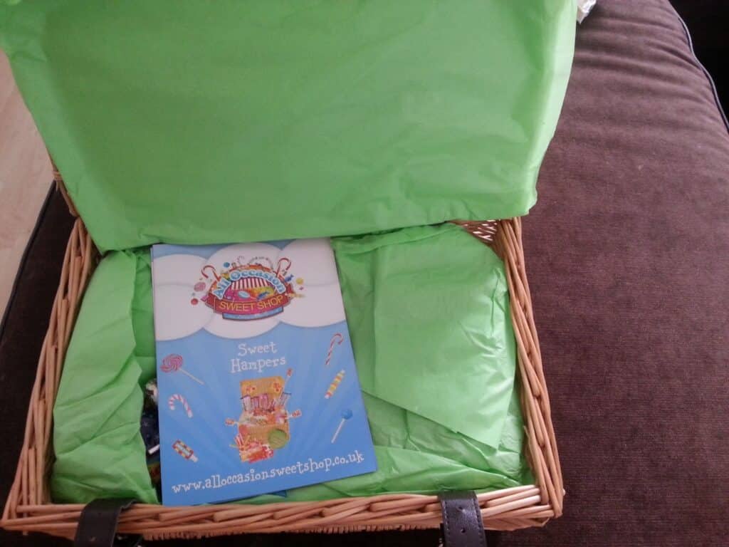 A wicker hamper with a leather strap is open on a brown surface. Inside, bright green tissue paper partly covers a colourful leaflet labelled "Sweetshop Sweet Hampers" with sweet illustrations and the website www.alloccasionssweetshop.co.uk.