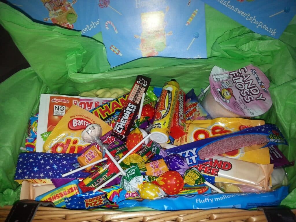 A wicker basket filled with assorted colourful sweets and chocolates, including lollipops, chewy bars, fizzy sweets, and chocolate, sitting on green tissue paper with a blue background inside the basket lid.