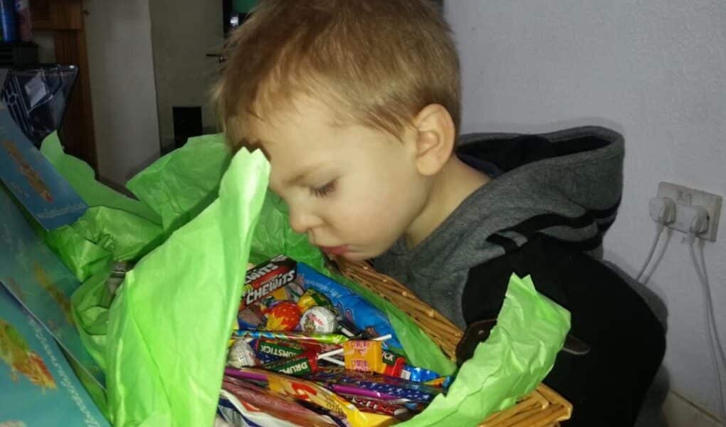 A young child with short light brown hair leans over a wicker basket filled with various colourful sweets. Green tissue paper lines the basket. The child appears focused on the sweets, with their face close to the basket. The setting is indoors.