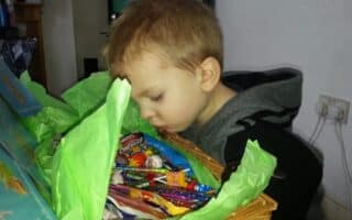 A young child with short light brown hair leans over a wicker basket filled with various colourful sweets. Green tissue paper lines the basket. The child appears focused on the sweets, with their face close to the basket. The setting is indoors.