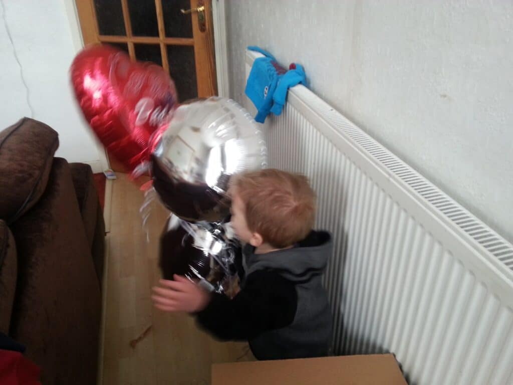 A small child with light brown hair faces a white radiator, holding shiny red and silver heart-shaped balloons. A blue toy sits on the radiator. In the background, a brown sofa and a wooden door with glass panes are visible.