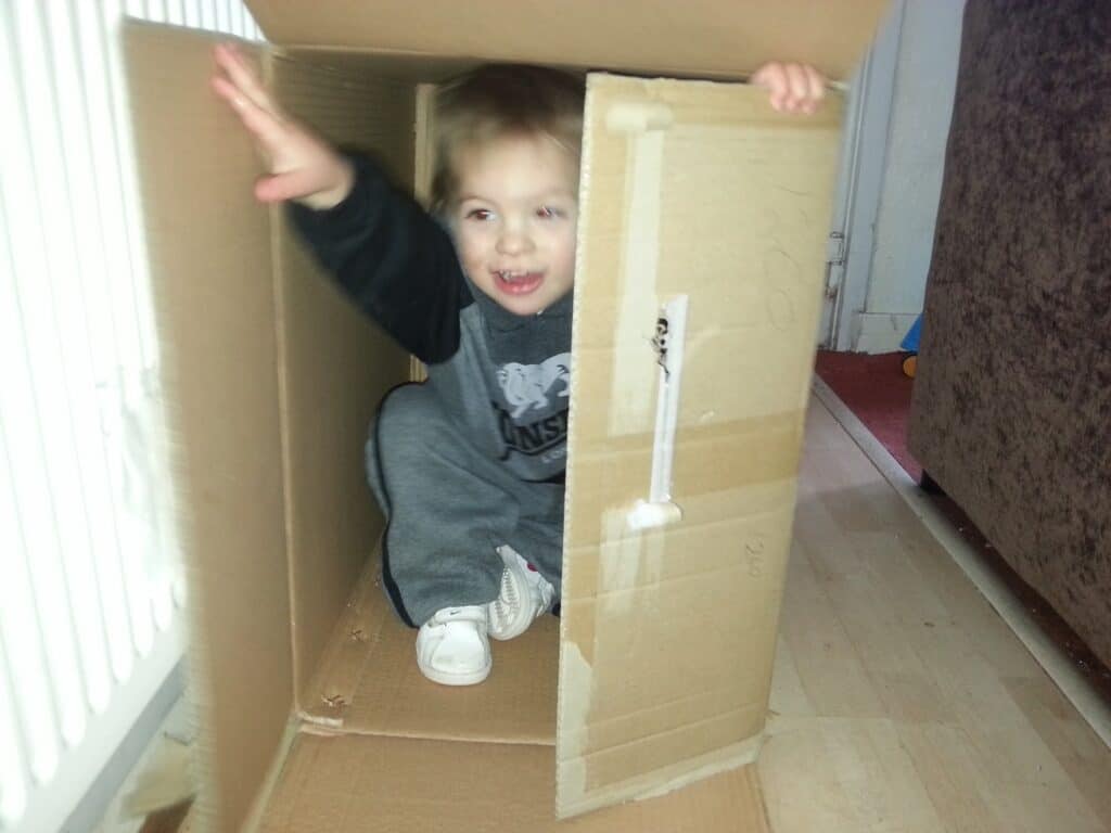 A young child smiles and waves while sitting inside a large cardboard box on a wooden floor. The box is open at both ends, and the child is dressed in a grey outfit with white shoes. Part of a white radiator and brown sofa are visible nearby.