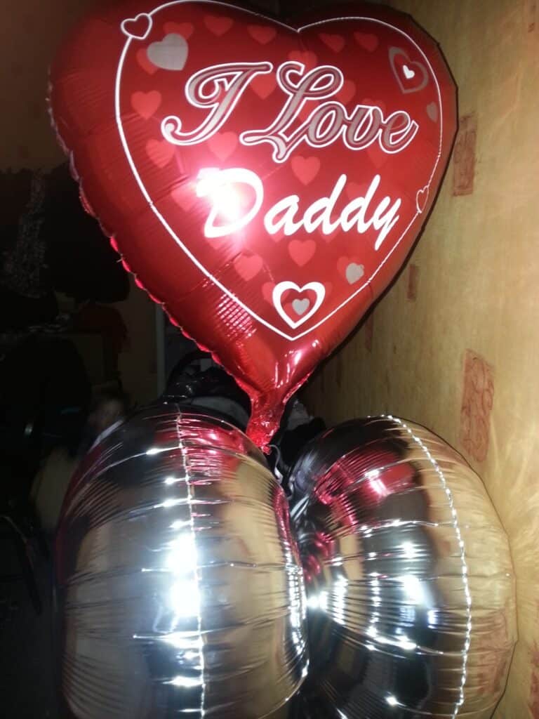 A heart-shaped red balloon with “I Love Daddy” written on it is tied to two silver balloons. The balloons are indoors against a yellowish wall with faded patterns. The silver balloons resemble a pair of buttocks from this angle.